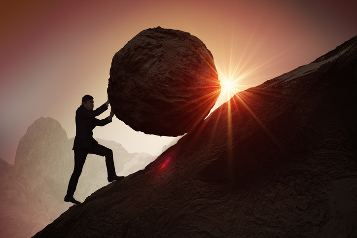 Silhouette of a businessman pushing a boulder up a hill.